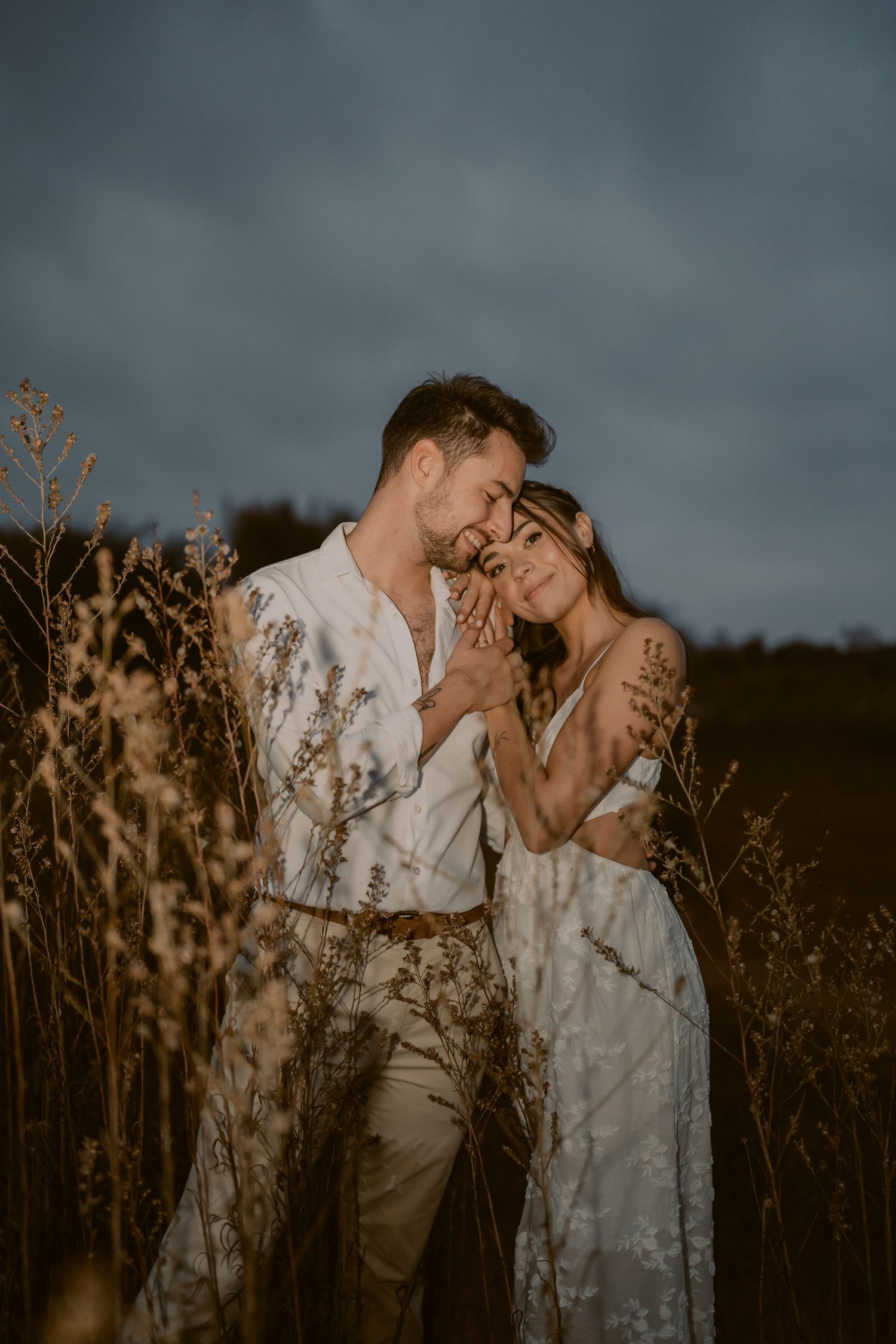 A romantic couple in white attire embraces tenderly among tall golden grasses under a dramatic dusk sky.