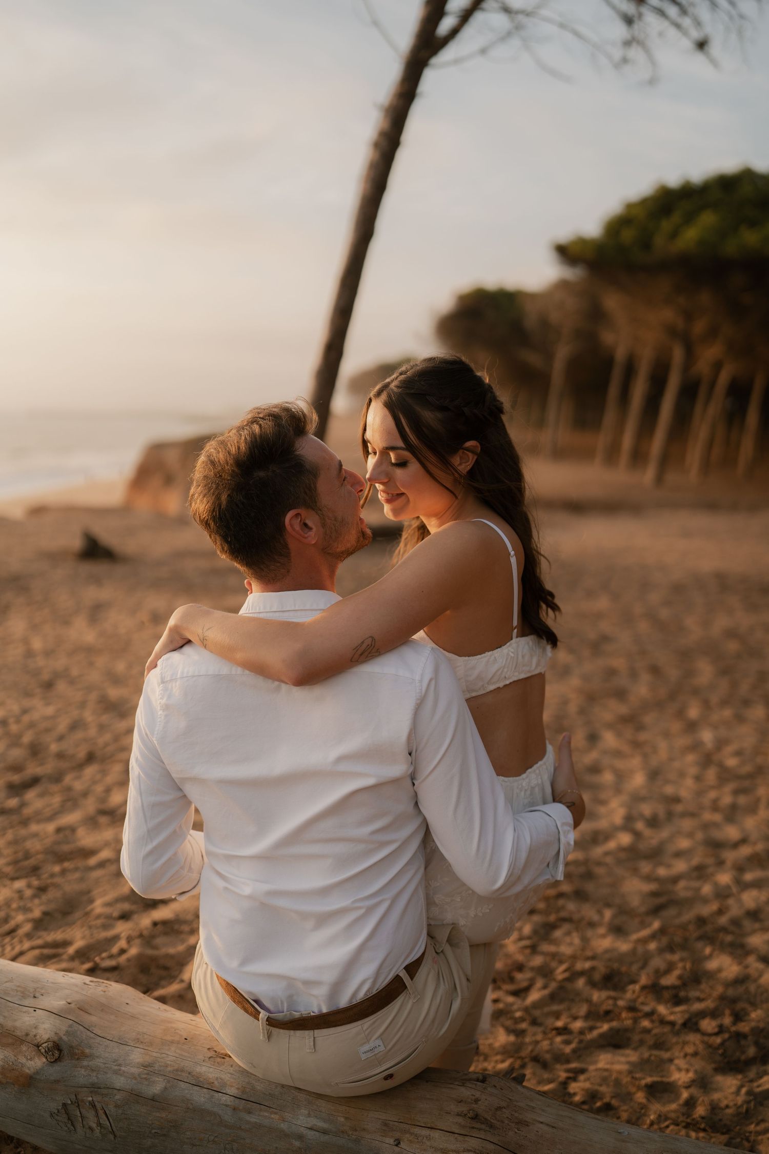 Couple in white outfits sharing a romantic moment on a sandy beach at golden sunset.