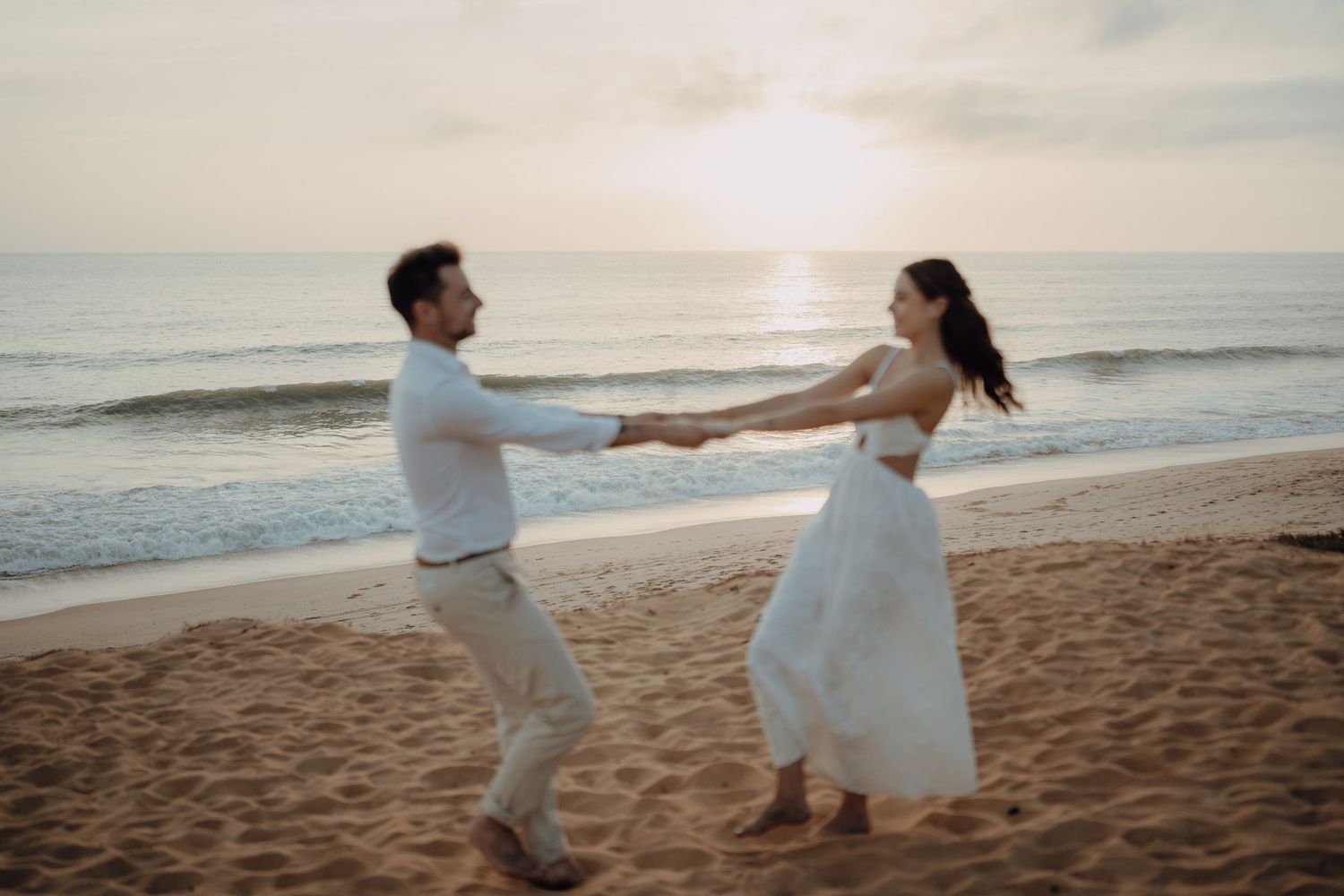 Couple dancing romantically on a sandy beach at sunset, holding hands in wedding attire by the ocean waves.