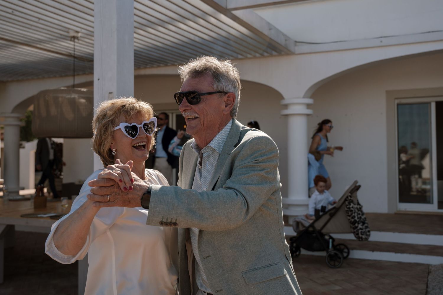 Elderly couple dancing together at outdoor wedding reception under white pergola in sunlight.
