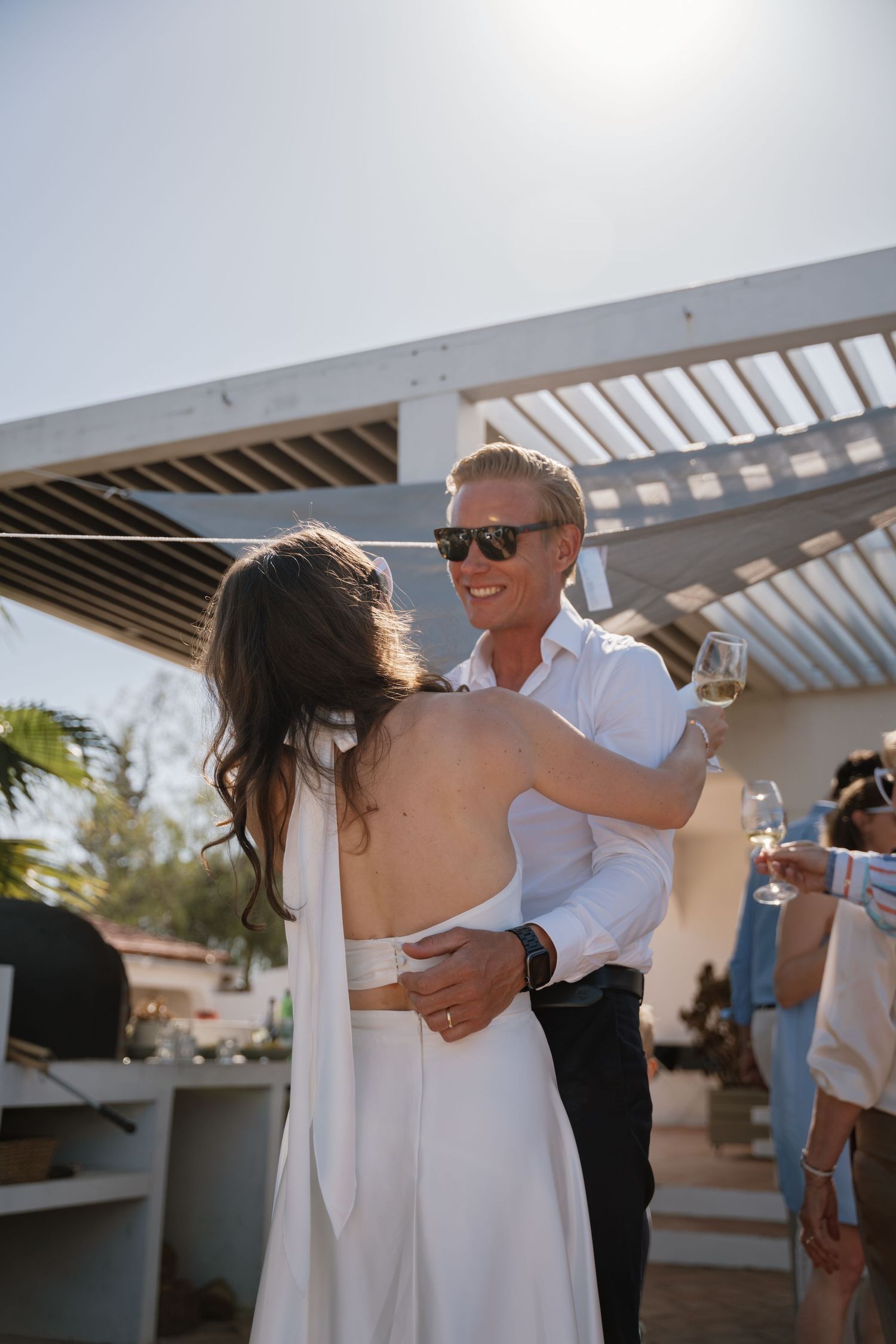 Groom in white shirt holding champagne embracing bride in backless white dress at sunny wedding.
