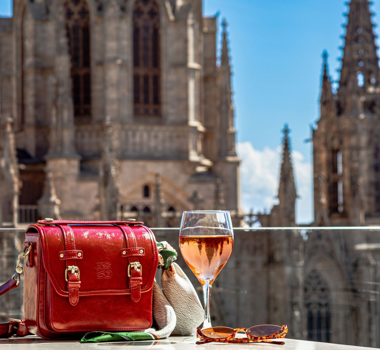 Red leather bag and rose wine glass on a sunny terrace overlooking Barcelona's Gothic Cathedral.