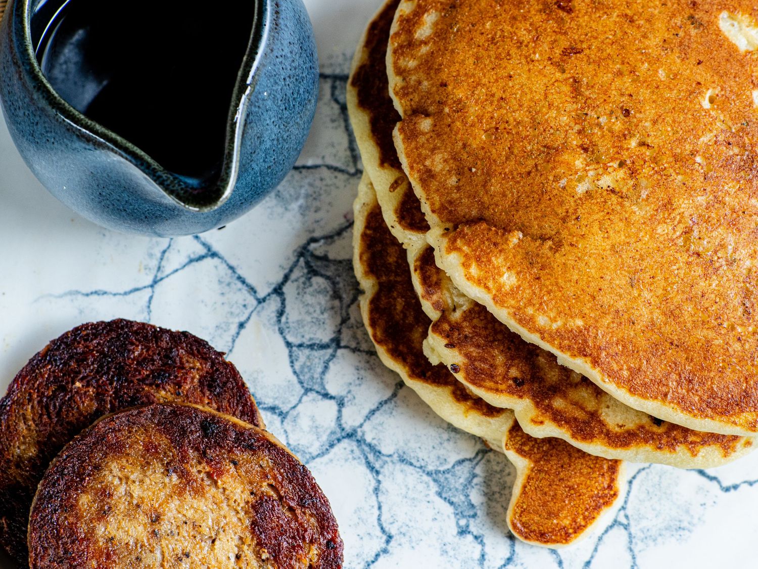 Stack of golden-brown pancakes with sausage patties and a blue ceramic pitcher of maple syrup on a marble surface.