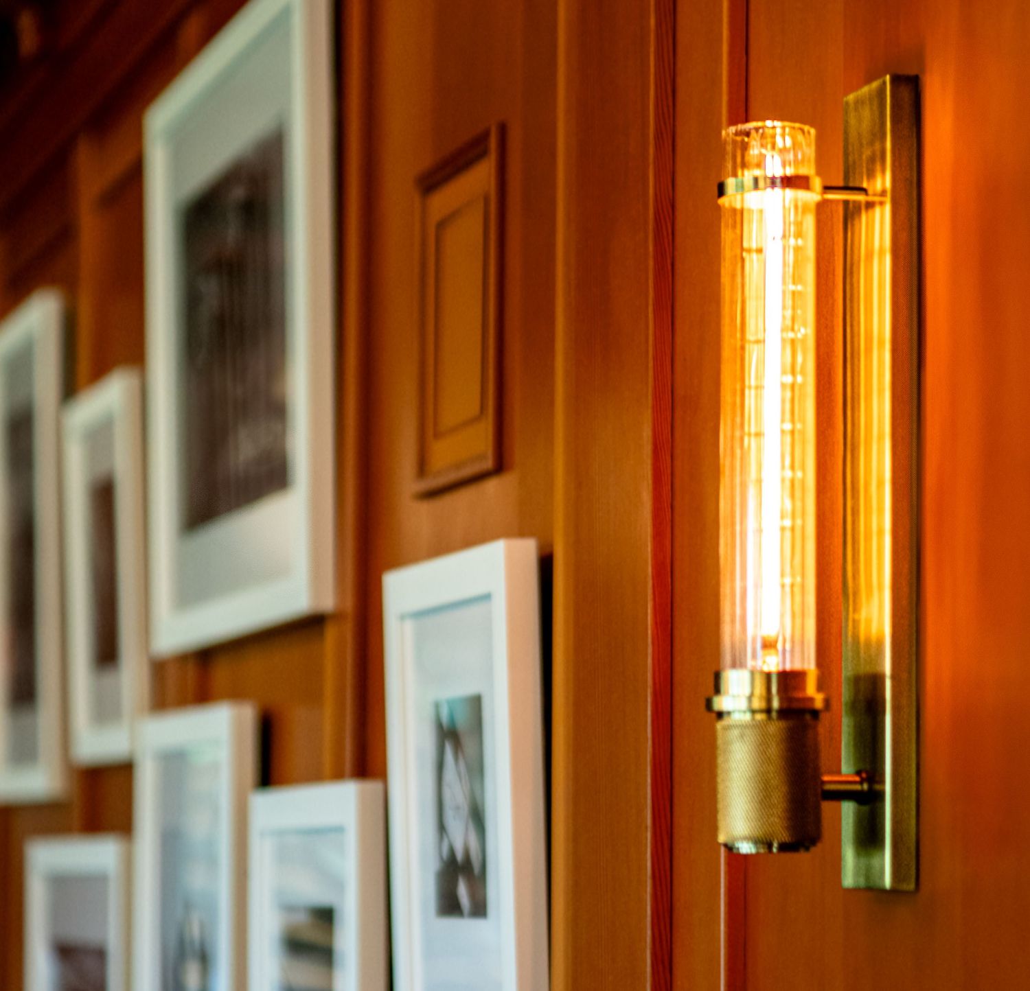Vintage Edison bulb wall sconce on warm wood-paneled wall with framed artwork gallery.