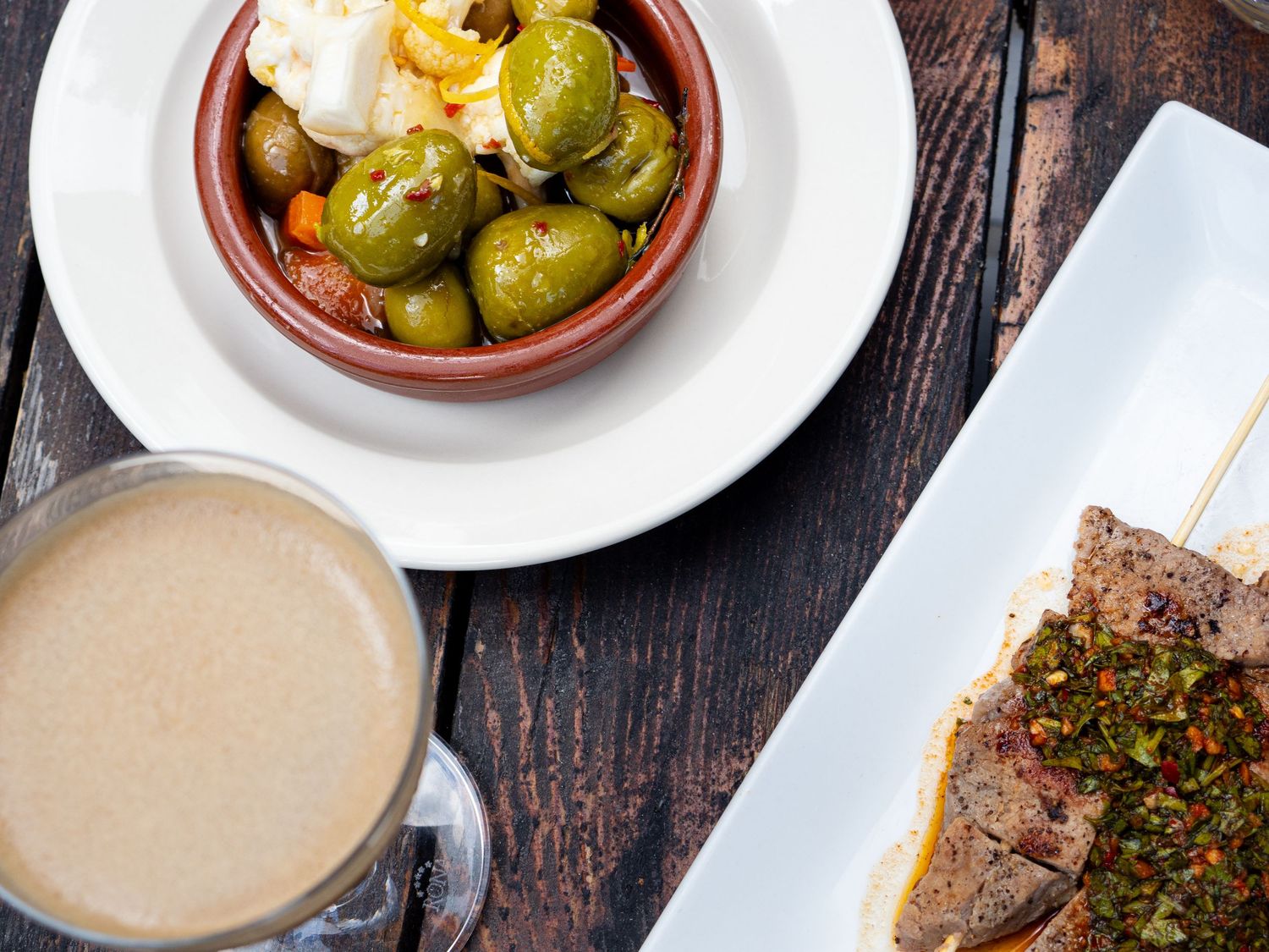 Overhead view of marinated olives, creamy hummus, and sliced meat with chimichurri on a rustic wooden table.