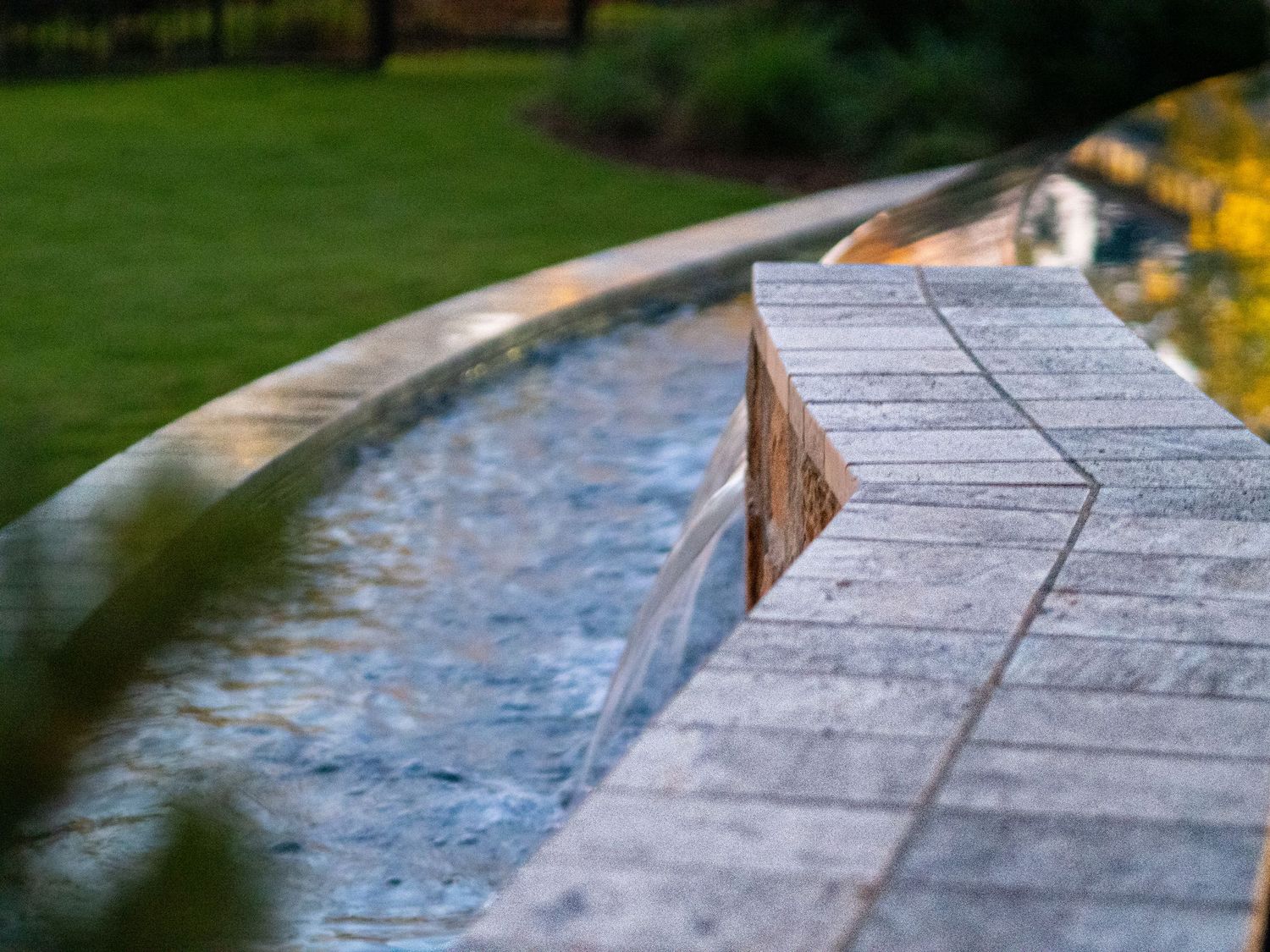 Close-up of a stone-edged water feature with a small waterfall cascading over granite tiles in a lush garden setting.
