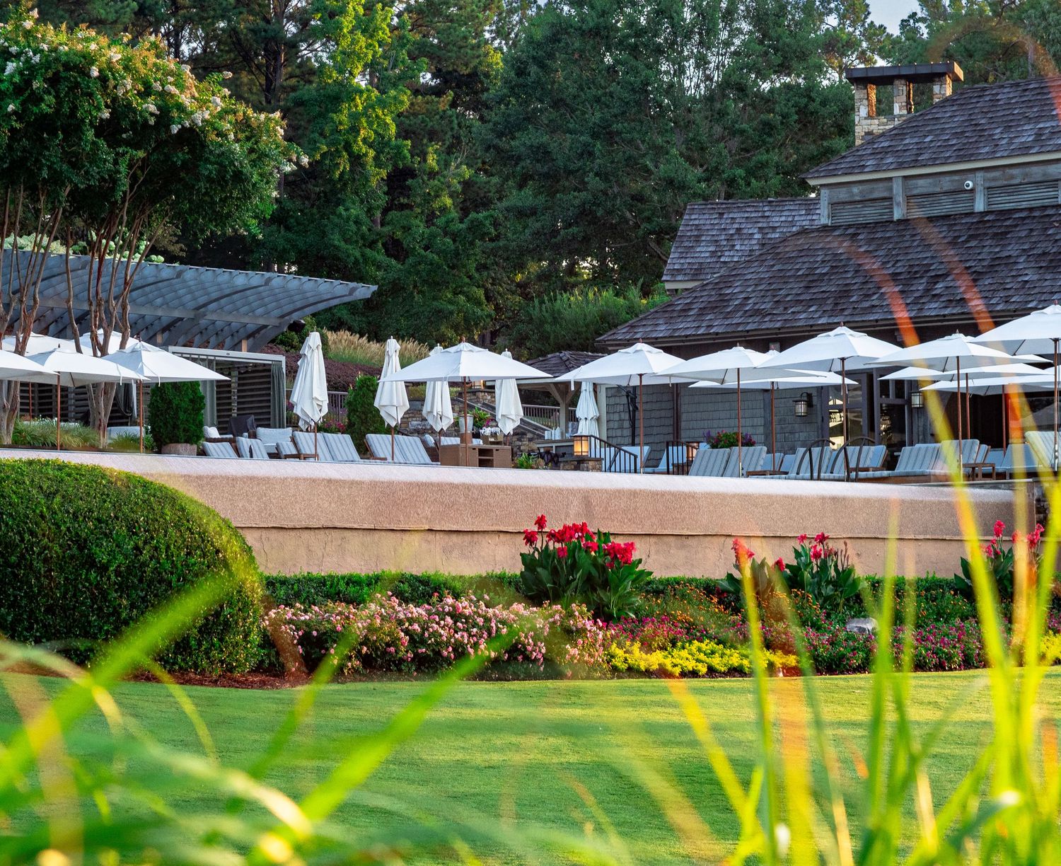 Elegant outdoor patio with white umbrellas, lounge chairs, and colorful garden beds beside a stone building.