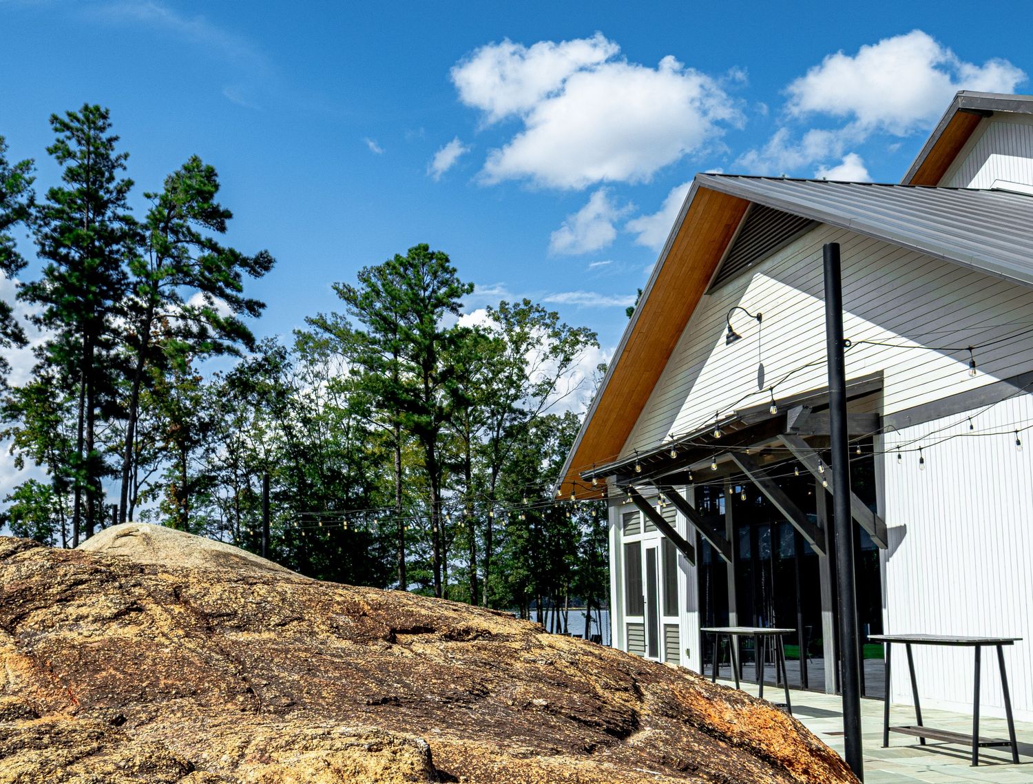 A modern white farmhouse-style building with metal roof nestled among tall pine trees beside a large granite rock outcropping.