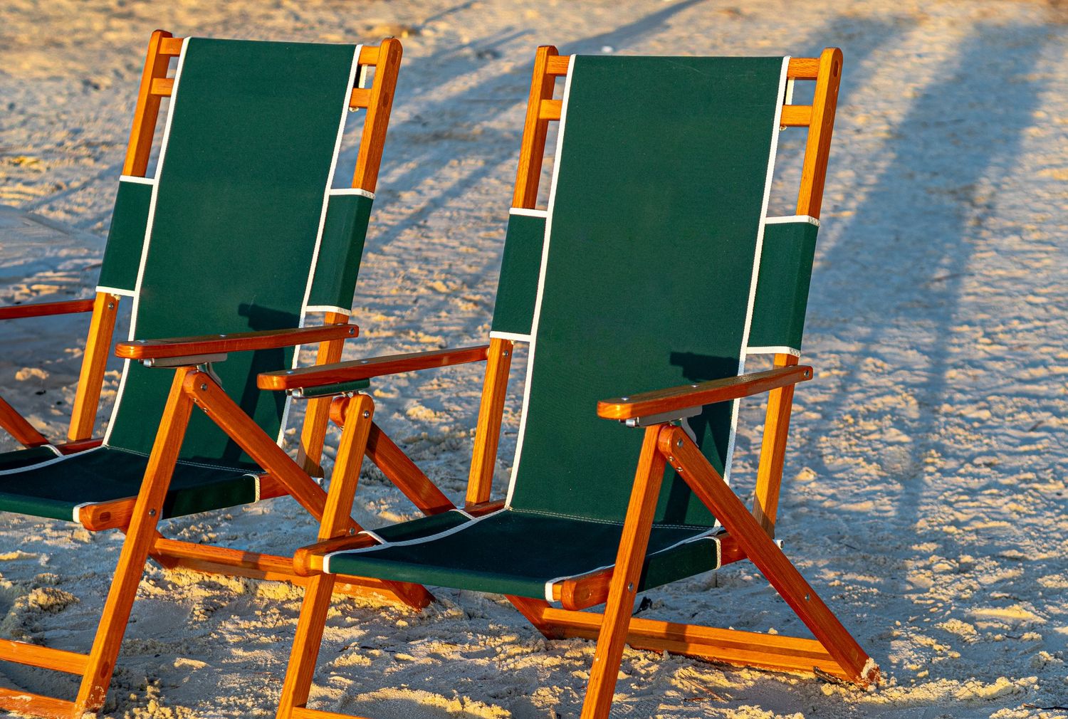 Two green wooden beach chairs casting long shadows on white sandy beach at golden hour.