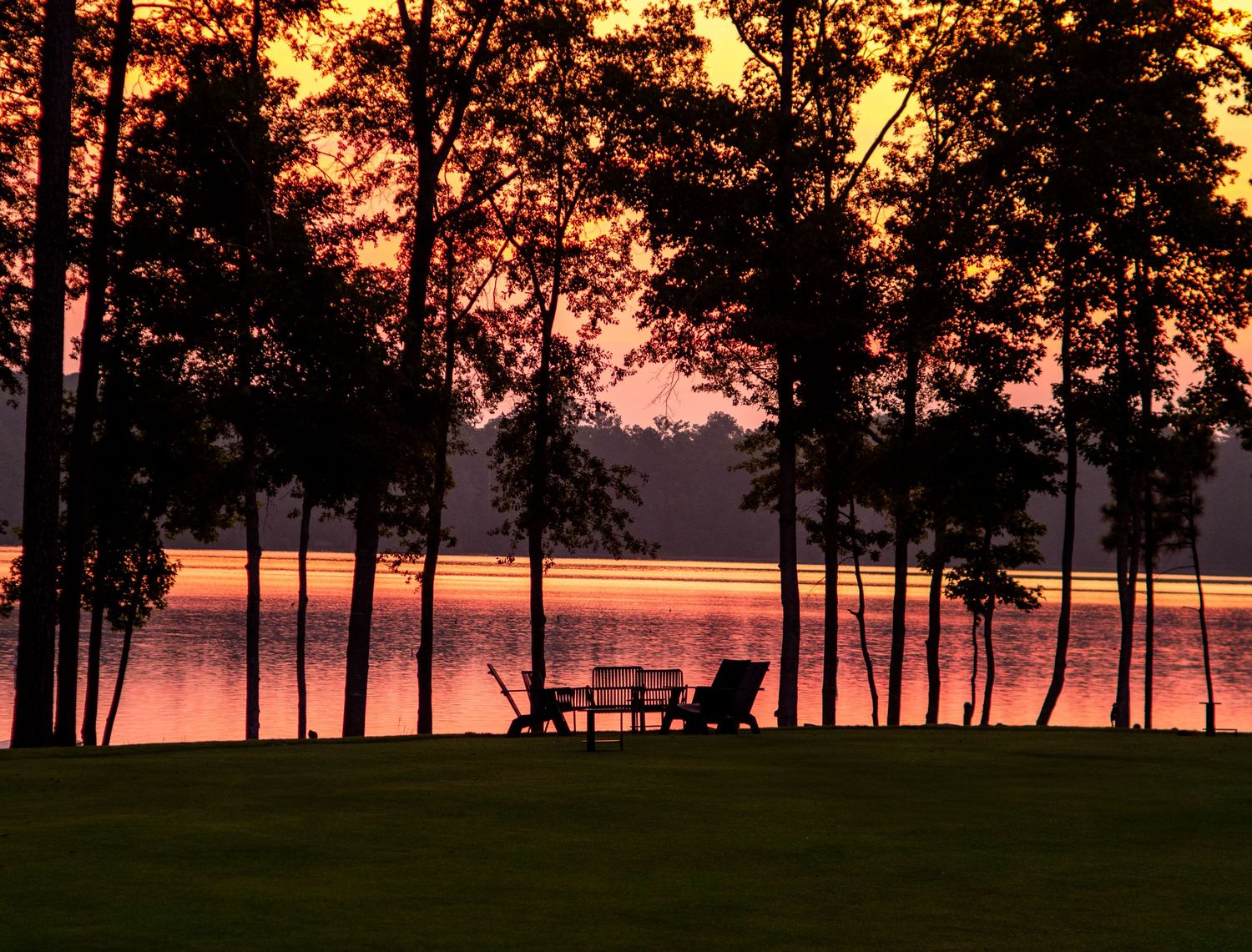 Silhouette of chairs by a lake at sunset with tall trees framing a vibrant orange and red sky.
