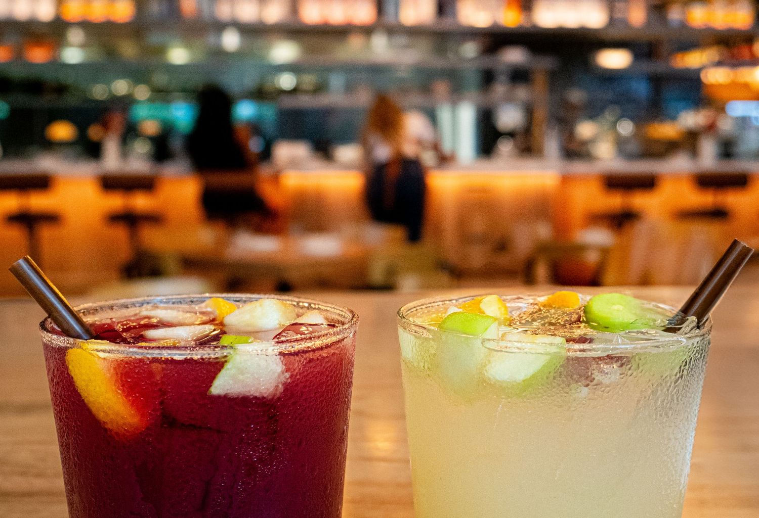 Two vibrant cocktails with metal straws and fresh fruit garnishes on a wooden bar counter in a warmly lit restaurant photographe montpellier