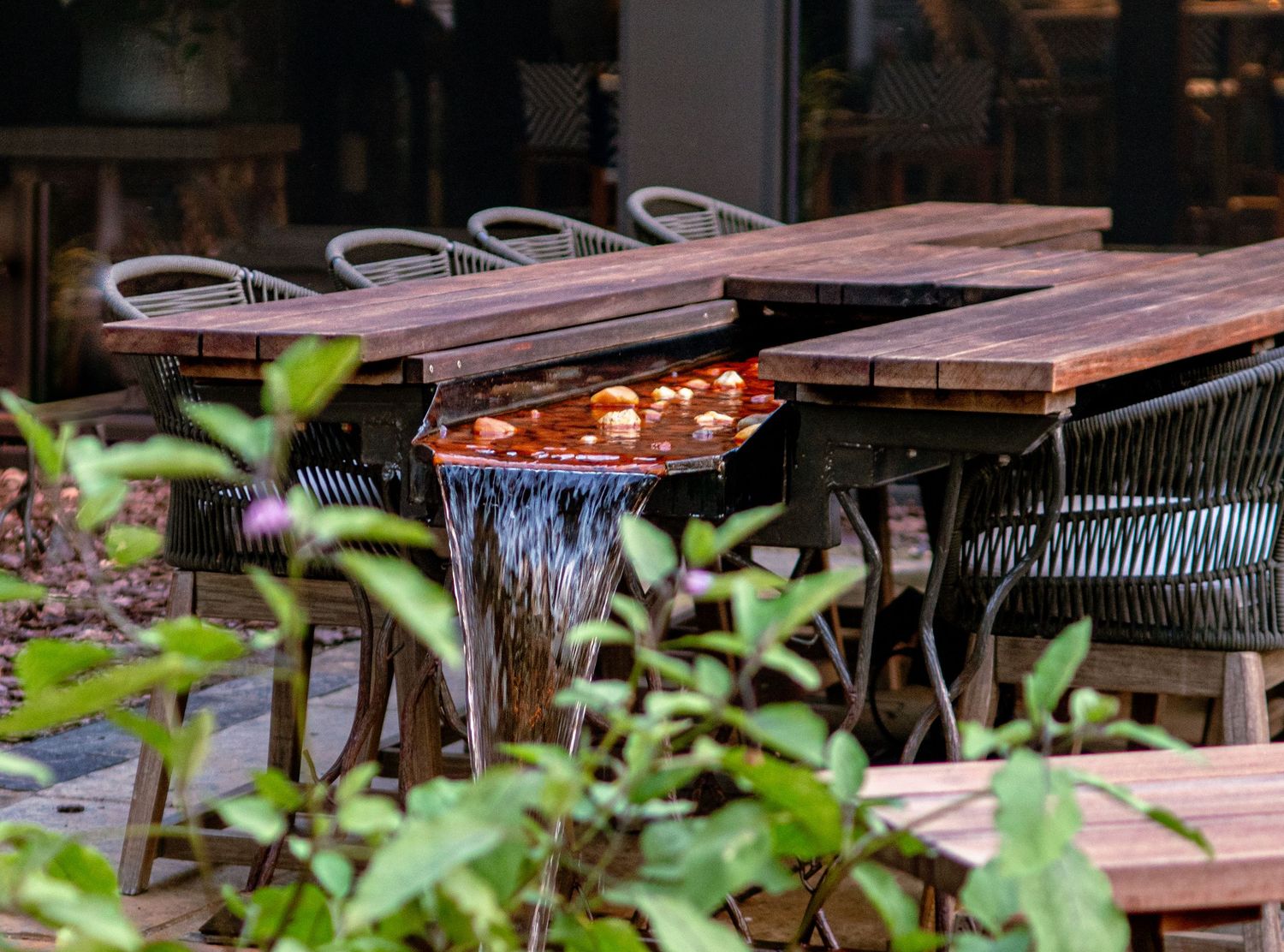 Rustic wooden outdoor dining tables with a small waterfall feature surrounded by lush green plants.