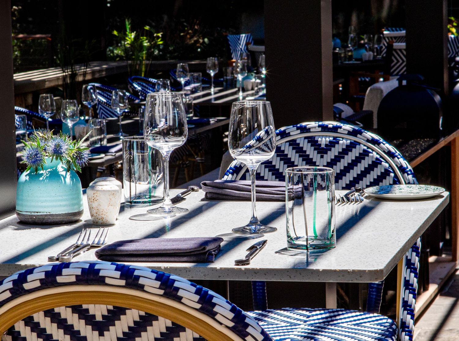 Elegant outdoor restaurant table set with blue and white woven chairs, crystal glasses, and a teal vase centerpiece. Hotel photographe