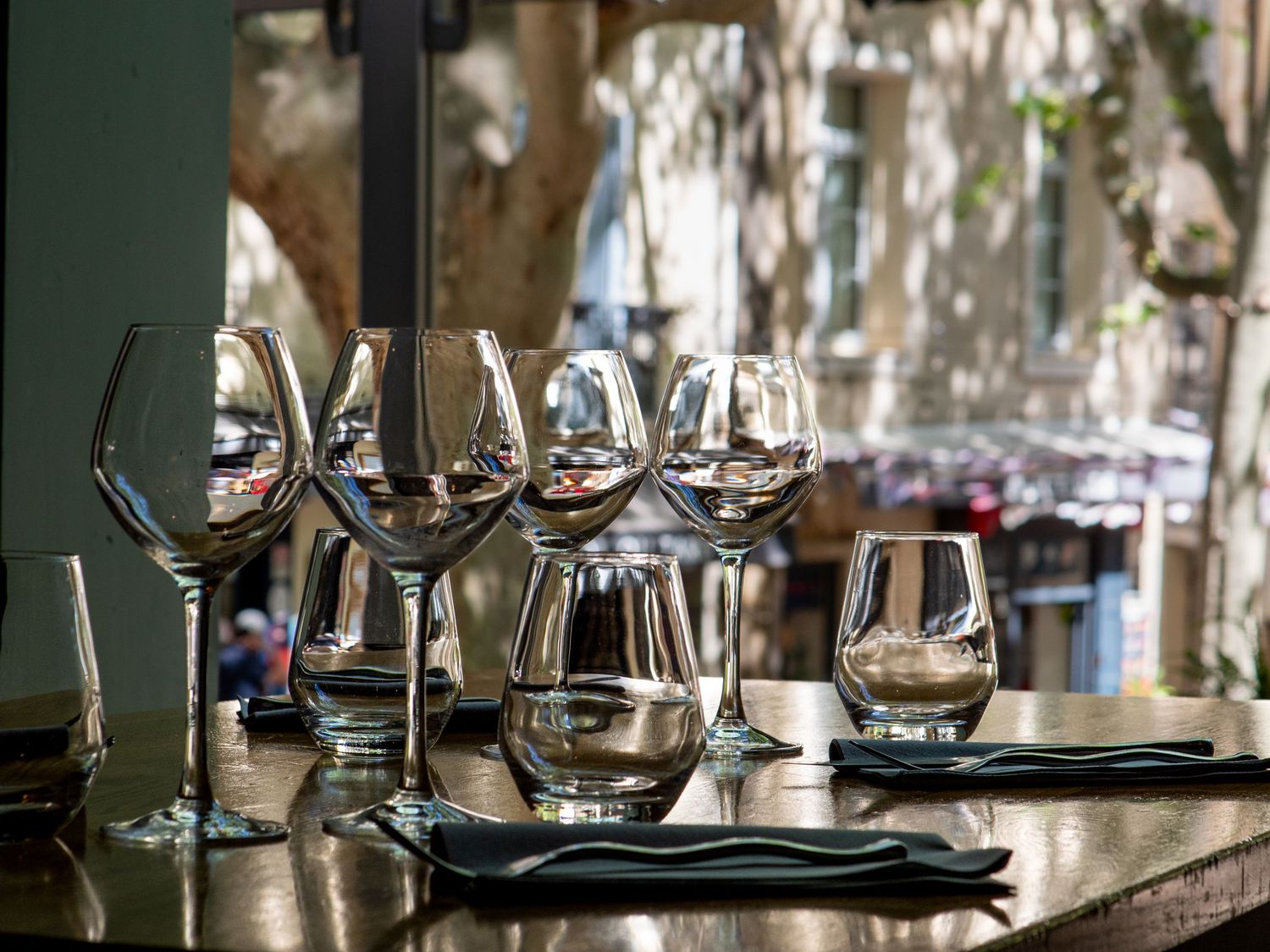 Empty wine glasses arranged on a wooden bar table at an outdoor restaurant patio with trees in the background. Sud de France photographe