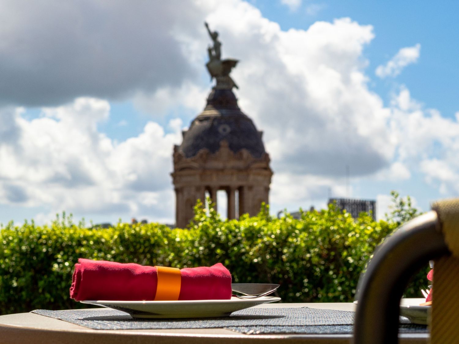 Red and yellow folded napkin on a rooftop terrace table with a monument statue and cloudy sky.