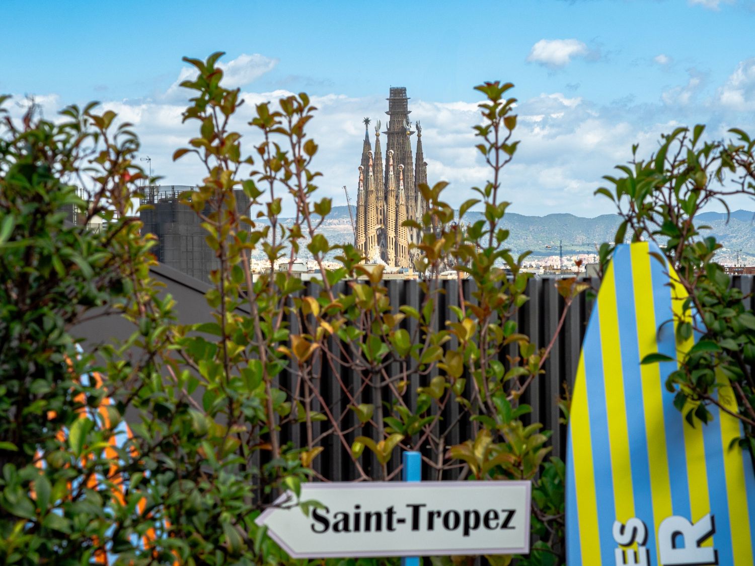 A Saint-Tropez directional sign with a surfboard beside lush green plants and the Sagrada Familia in the background.