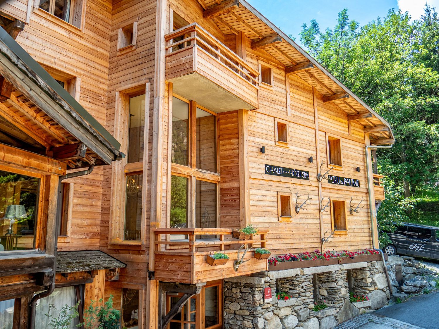 Rustic wooden alpine chalet with balconies and stone foundation surrounded by lush green trees in a mountain setting.