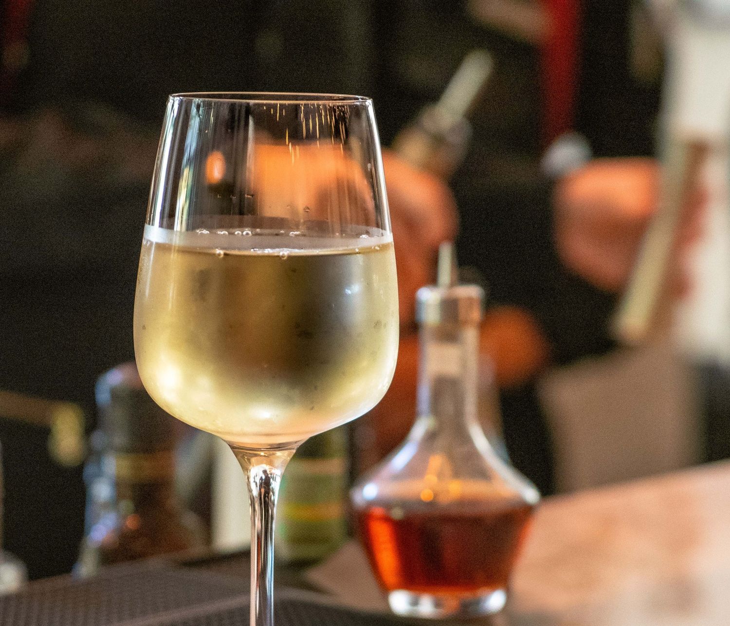 Close-up of a white wine glass with a blurred bartender and decanter in the background.