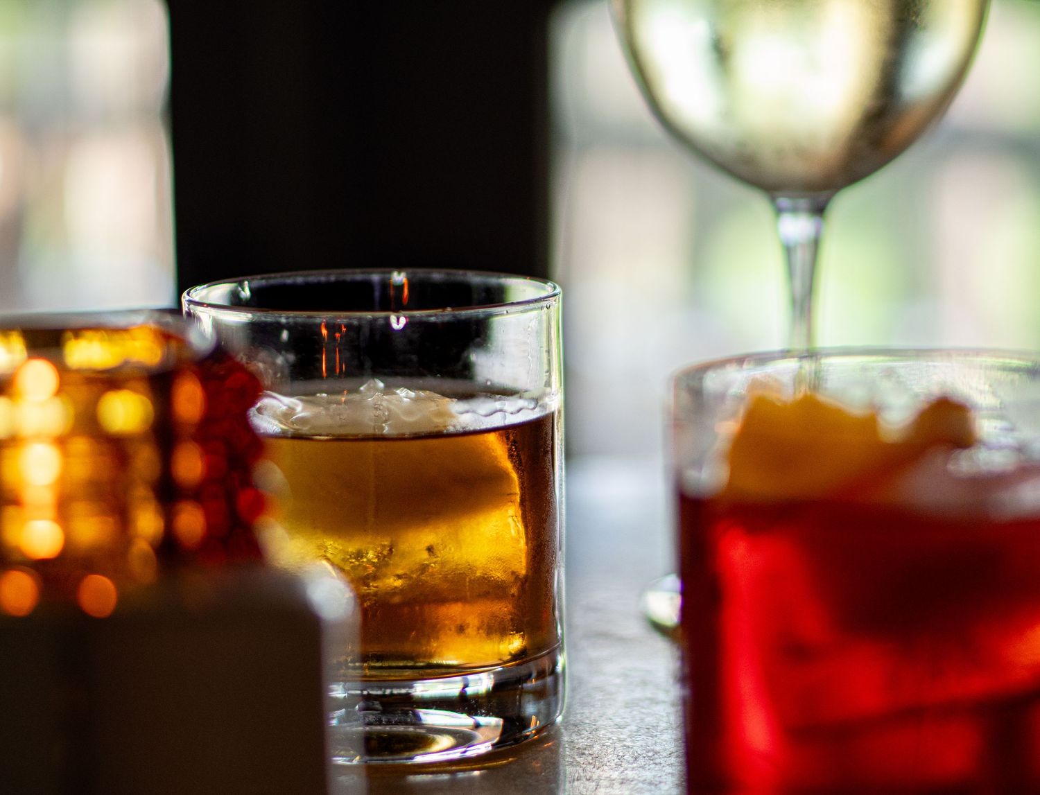 Amber whiskey cocktail with ice sphere beside a red cocktail and wine glass on a dimly lit bar.