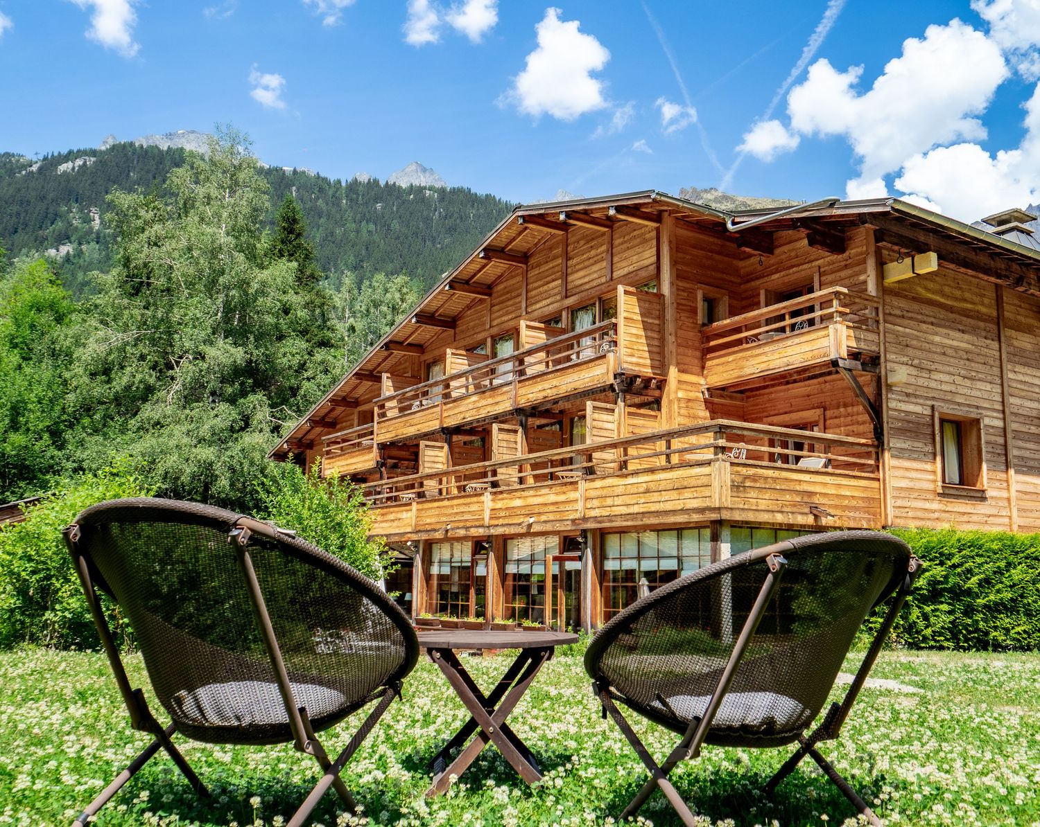 Wooden alpine chalet with balconies, two chairs on green meadow, mountain forest backdrop on sunny day.