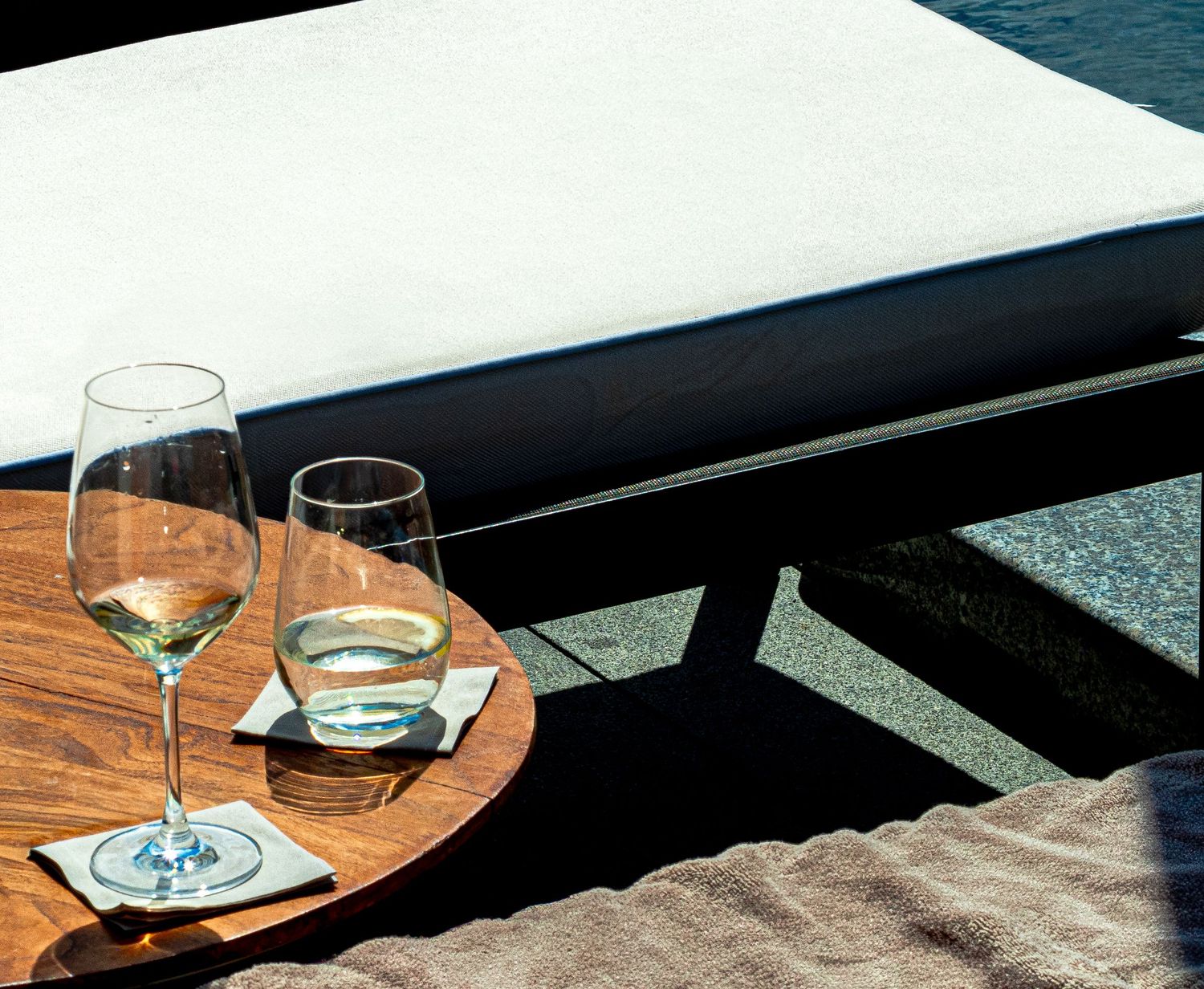 Two elegant wine glasses with white wine on a round wooden table beside a luxury poolside lounge chair. photographer hotel Spain