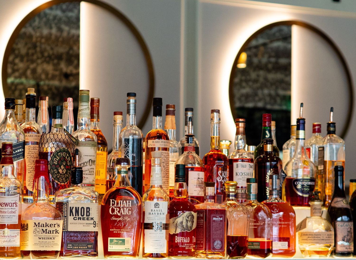 A large collection of premium whiskey and bourbon bottles displayed on a bar shelf, including Maker's Mark and Knob Creek.
