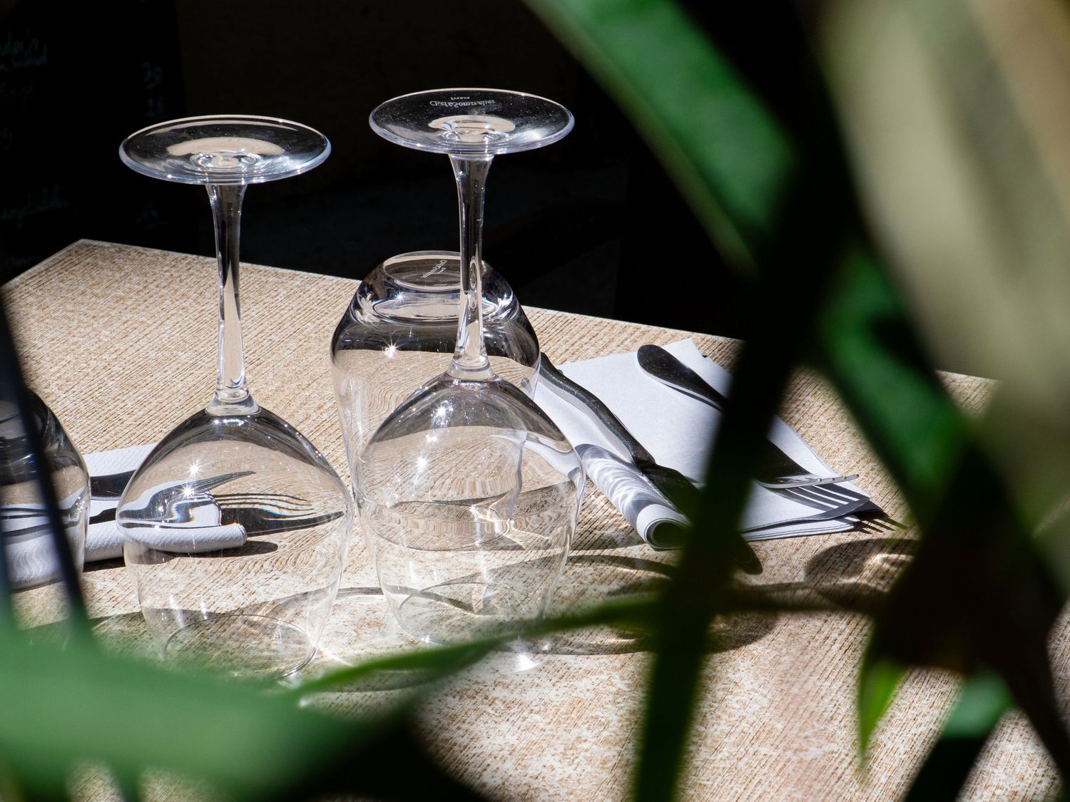 Two elegant wine glasses placed upside down on a table set with cutlery and linen, surrounded by lush green plants. Restaurant photographe