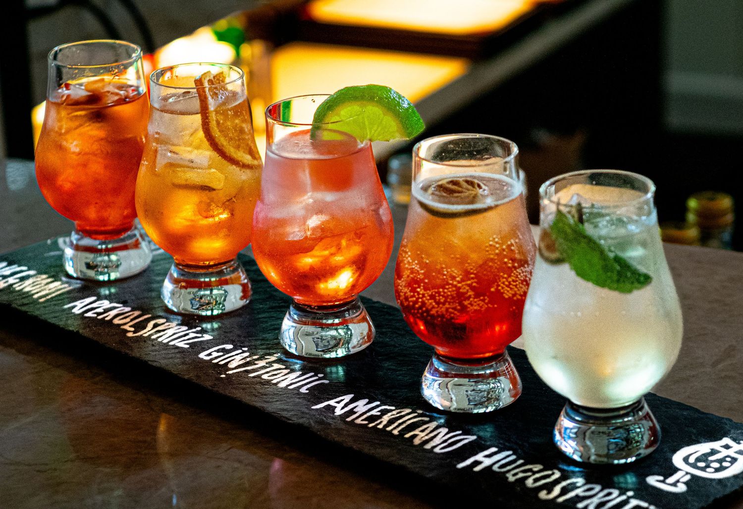 Flight of five colorful cocktails displayed on a branded wooden serving board at a bar.