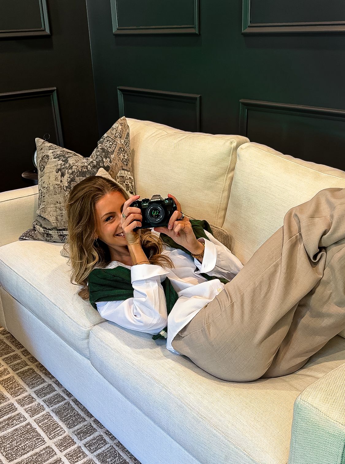 Woman lying on cream sofa holding a camera, wearing white shirt and green vest, with decorative pillows nearby.