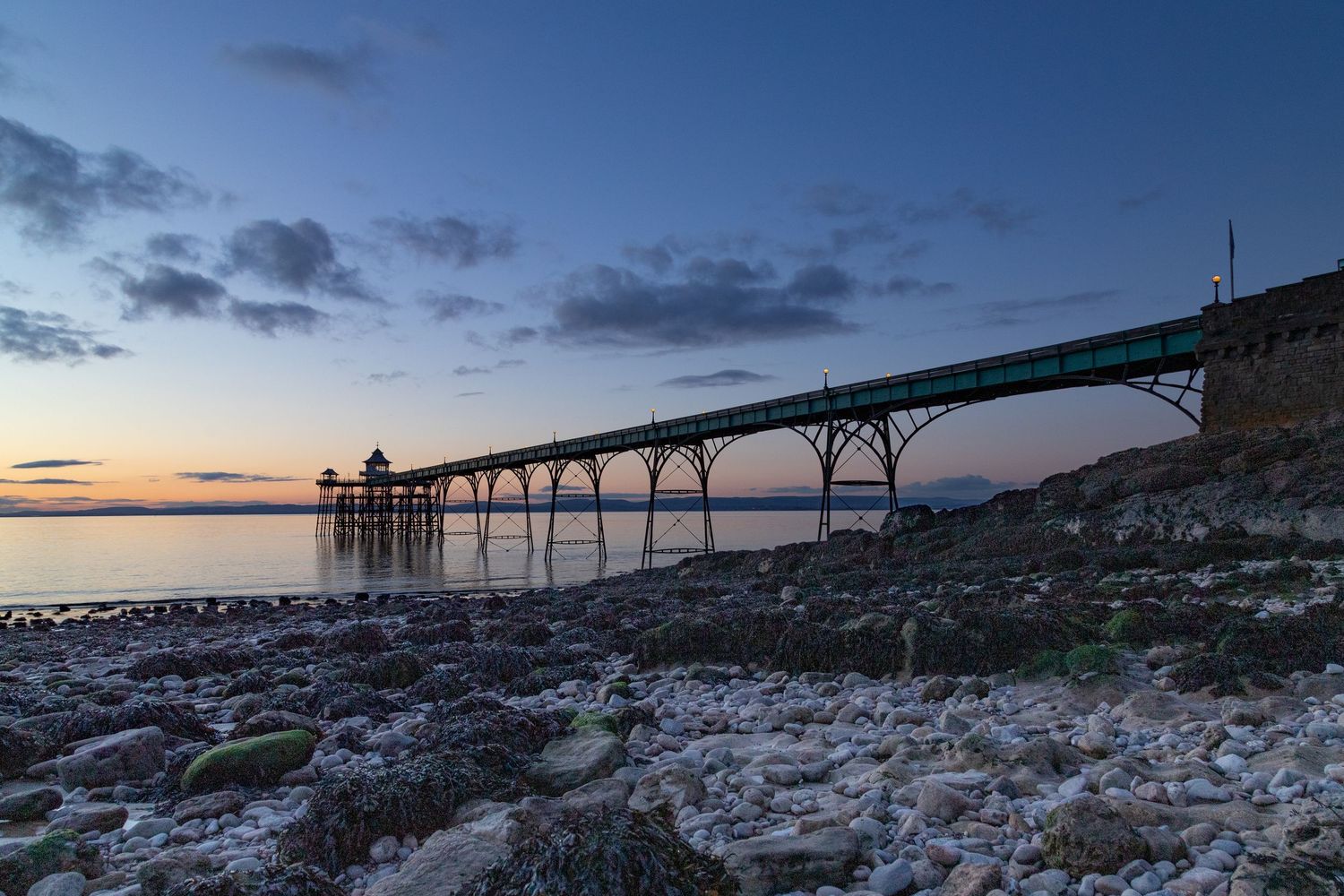 Clevedon Pier in Somerset at sunset with rocks in the foreground and calm sea.