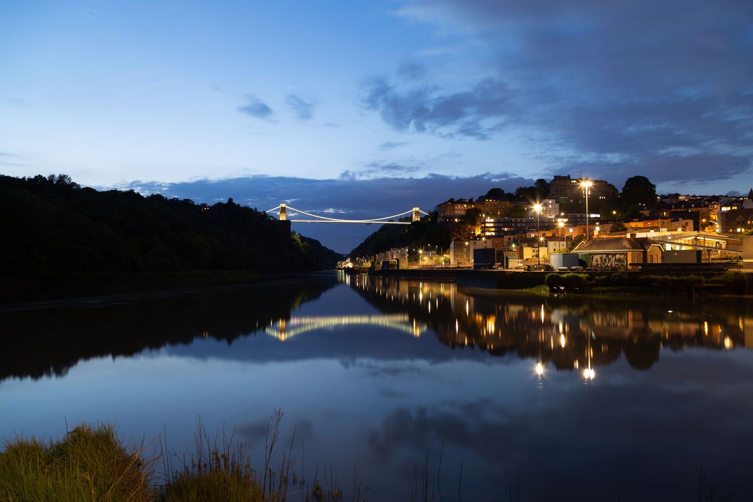 Clifton Suspension Bridge in Bristol at dusk with city lights reflected on the River Avon.