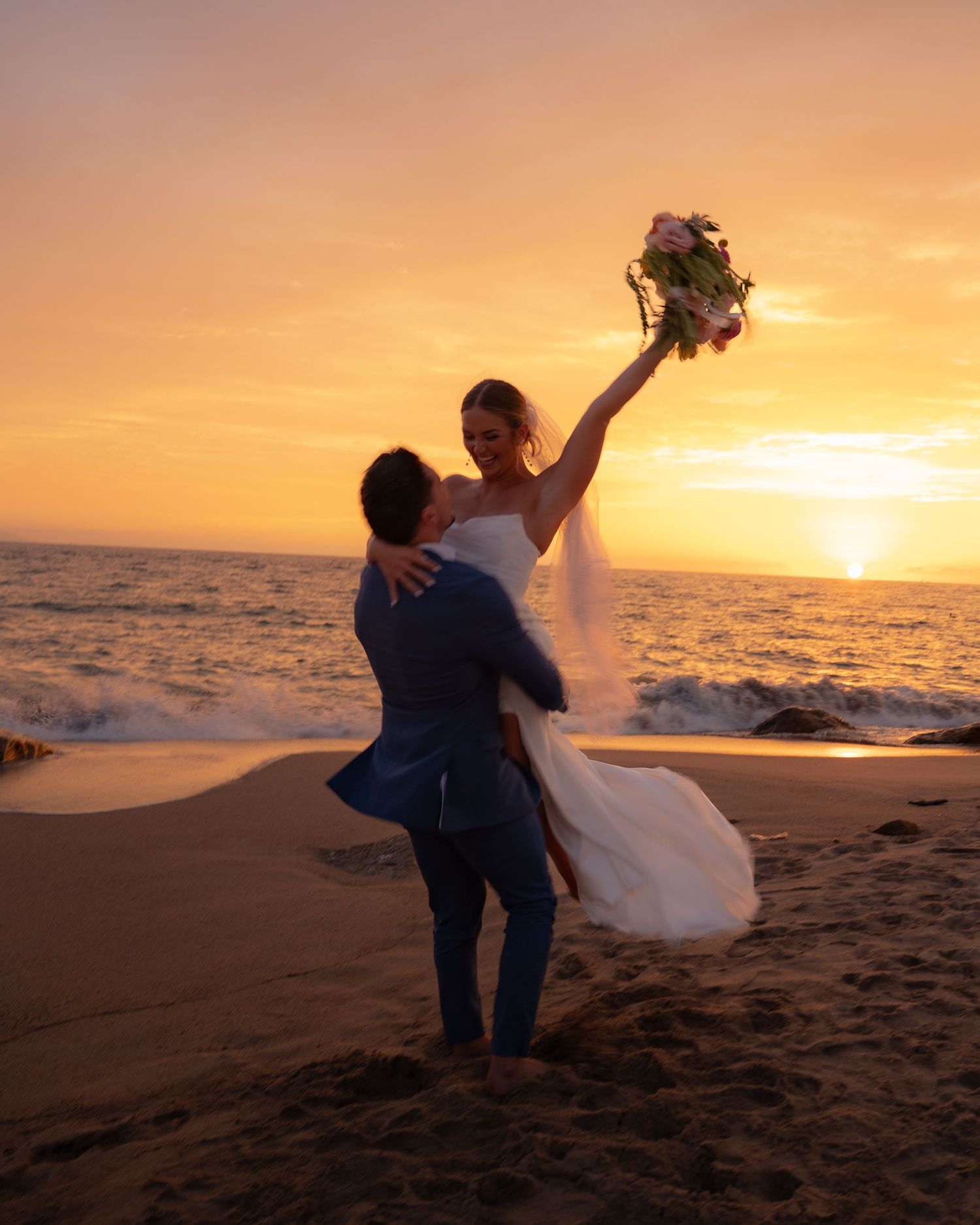 Groom lifts bride on a sandy beach as she raises her bouquet against a vibrant golden sunset.