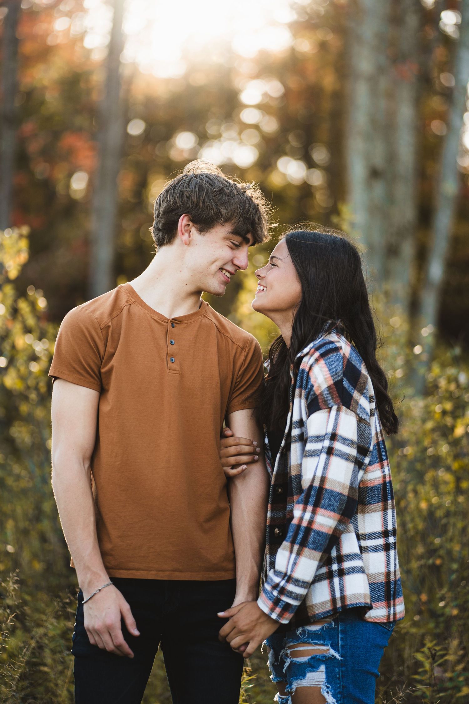 Young couple holding hands and smiling at each other in a sunlit autumn forest setting.