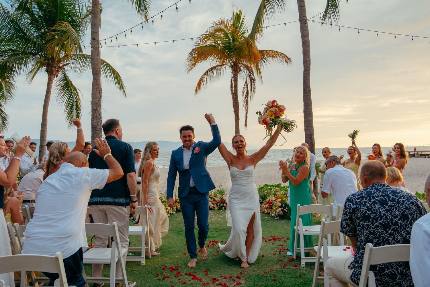 Joyful newlyweds raise arms celebrating as they walk down the aisle at tropical beachside wedding ceremony.