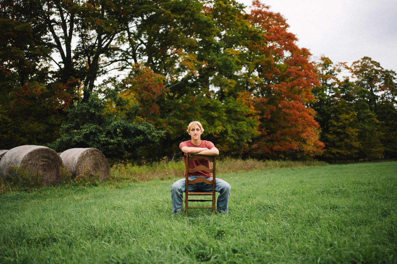 Young man sitting on a chair in a green field with colorful autumn trees and hay bales.