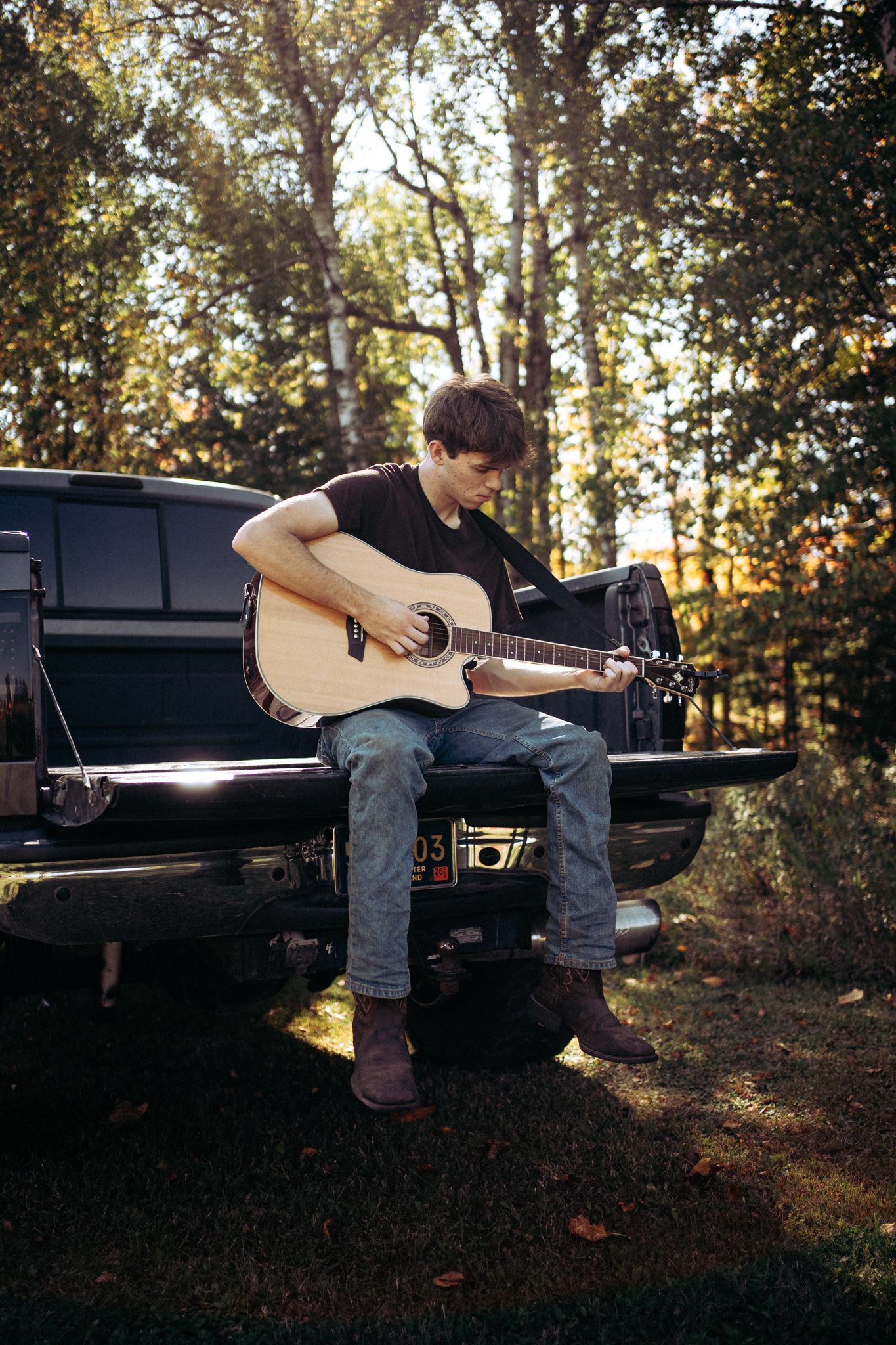 Young man playing acoustic guitar while sitting on a truck tailgate in a wooded setting.