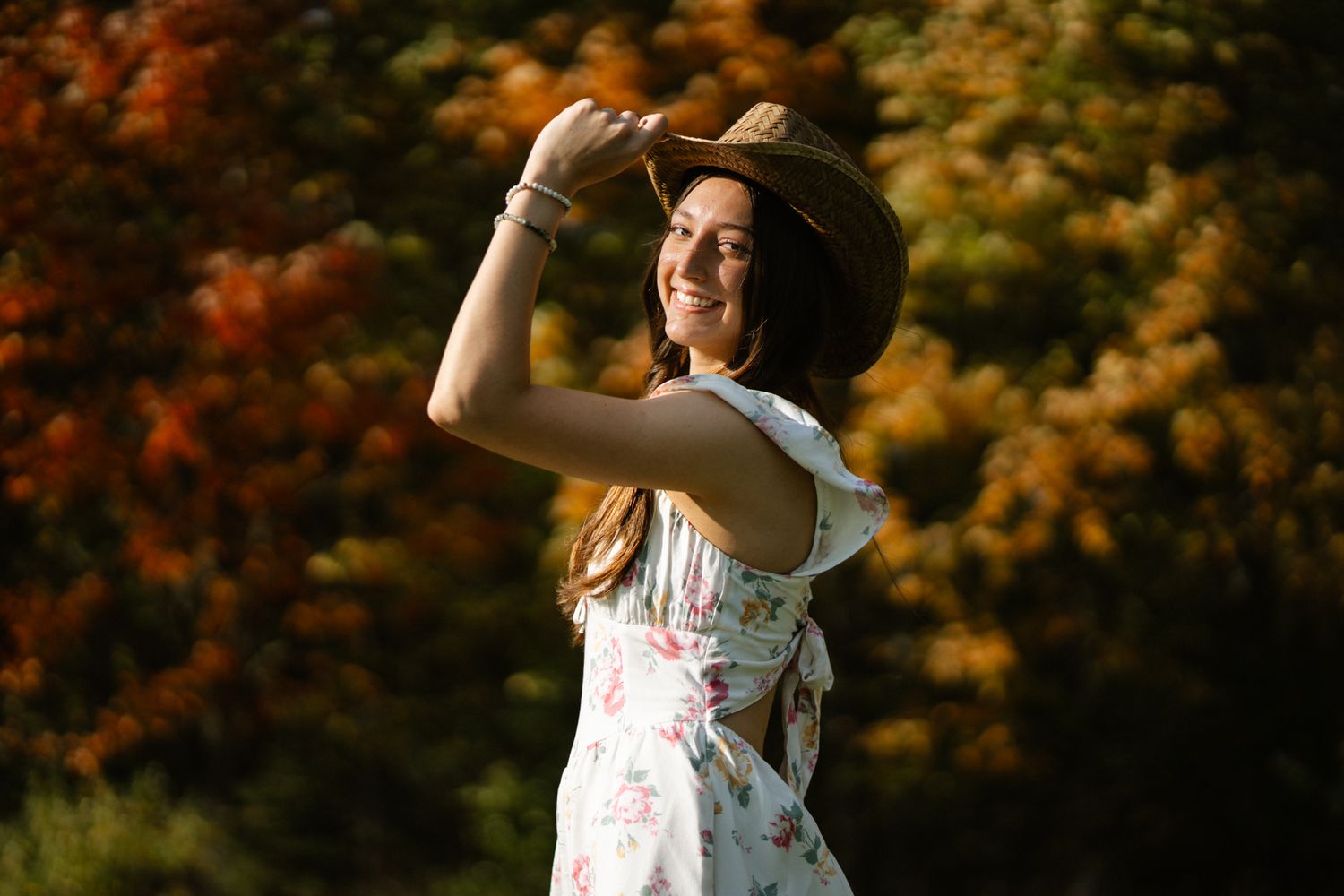 Smiling young woman in floral dress holding a wide-brim hat outdoors with colorful fall foliage.