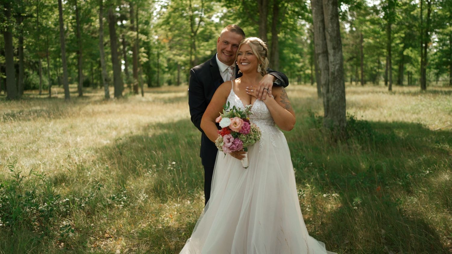 Bride and groom posing romantically in a sunlit forest, bride holding a colorful bouquet.