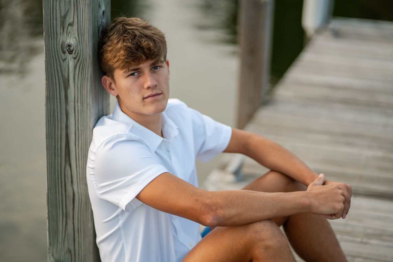 Teen boy in white polo shirt sitting on wooden dock leaning against post near calm water.