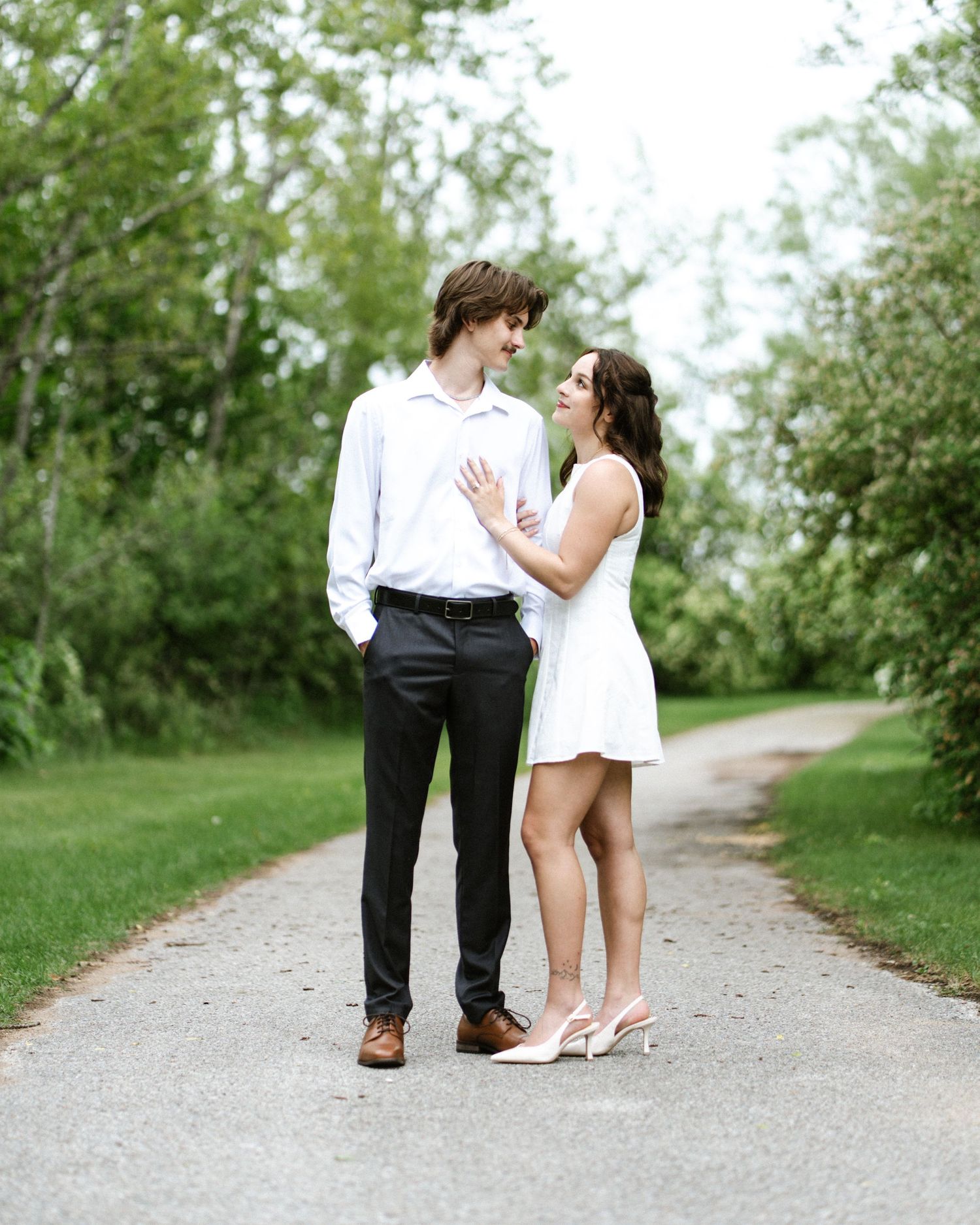 A couple in white outfits share a romantic moment on a tree-lined path during an outdoor engagement photo session.