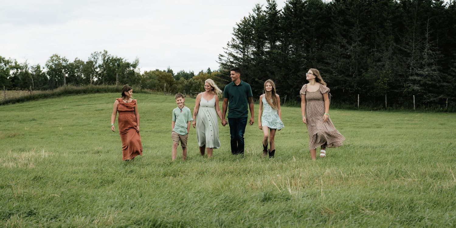 Family of six holding hands walking through open green field near pine tree line.