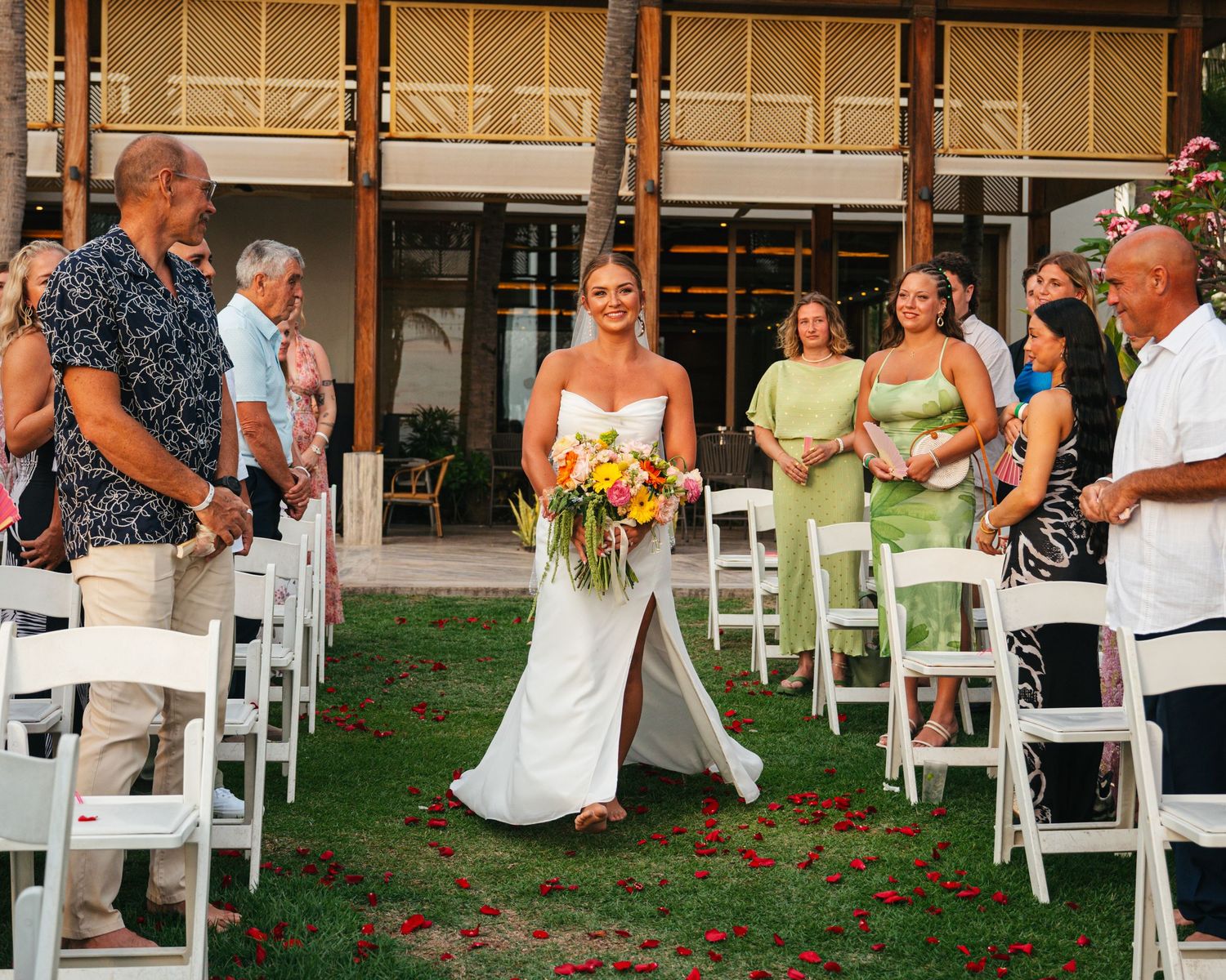 Radiant bride walks down a rose petal aisle at an outdoor tropical wedding ceremony with white chairs.