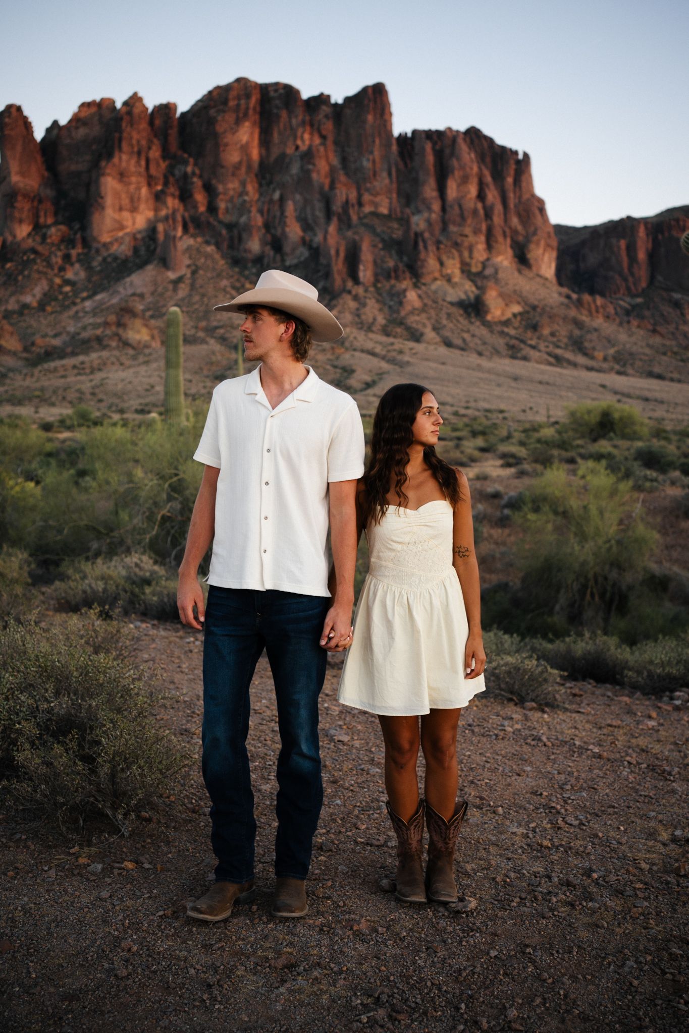 Couple in white outfits holding hands before dramatic desert mountain landscape at sunset.