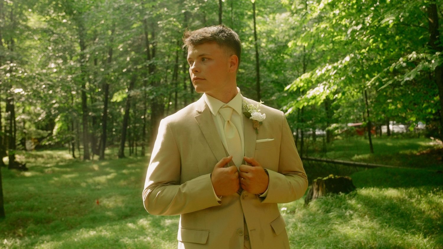 A groom in a tan suit adjusts his jacket while standing in a sunlit forest on his wedding day.