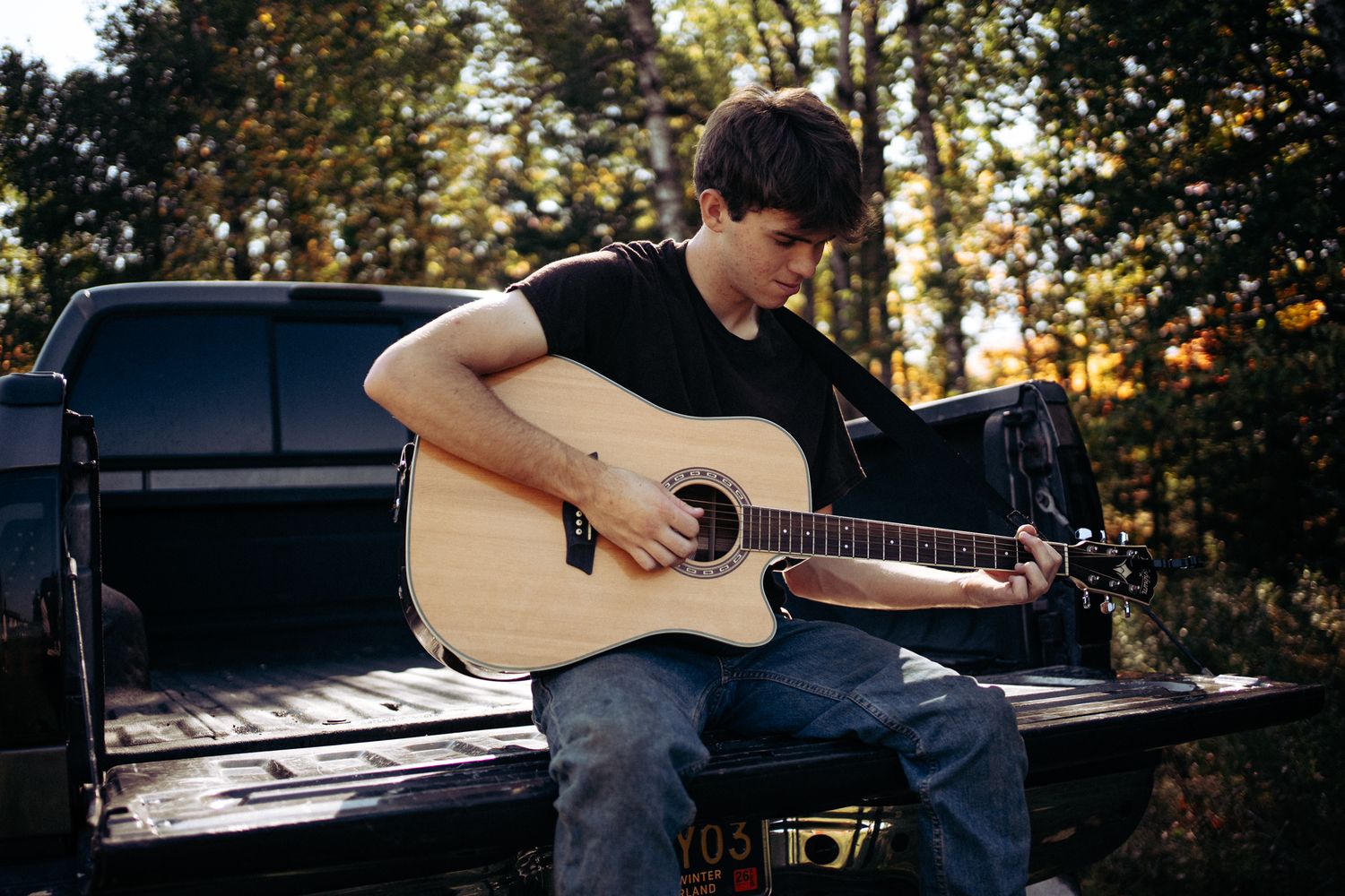Teenage boy playing acoustic guitar while sitting on truck tailgate at sunset in woods.