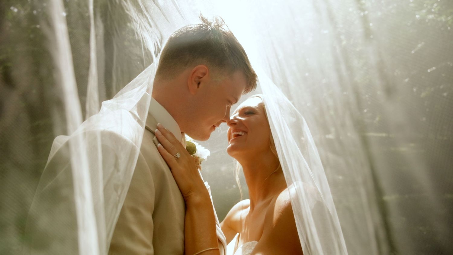 Bride and groom share romantic moment under sheer veil in soft sunlit outdoor setting.