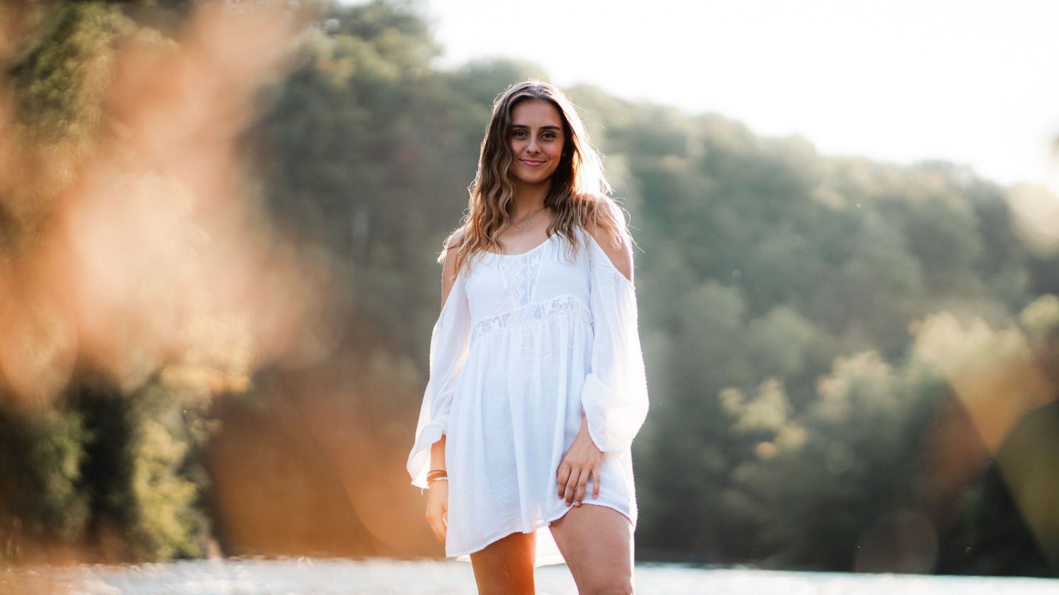 Smiling woman in white off-shoulder dress standing by a sunlit lake with trees in background.
