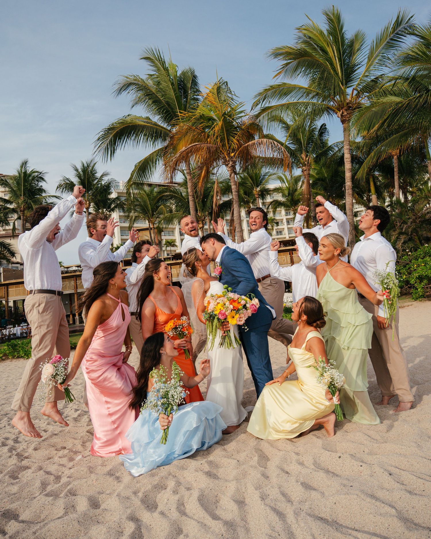 Wedding party celebrating with bride and groom kissing on a tropical beach with palm trees.