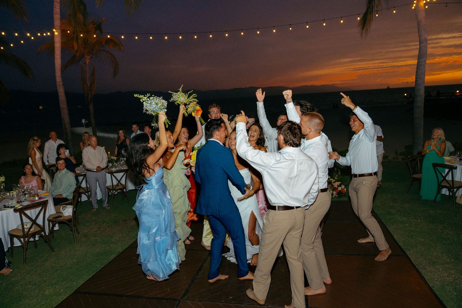 Wedding guests dance joyfully under string lights at outdoor tropical beach reception at sunset.