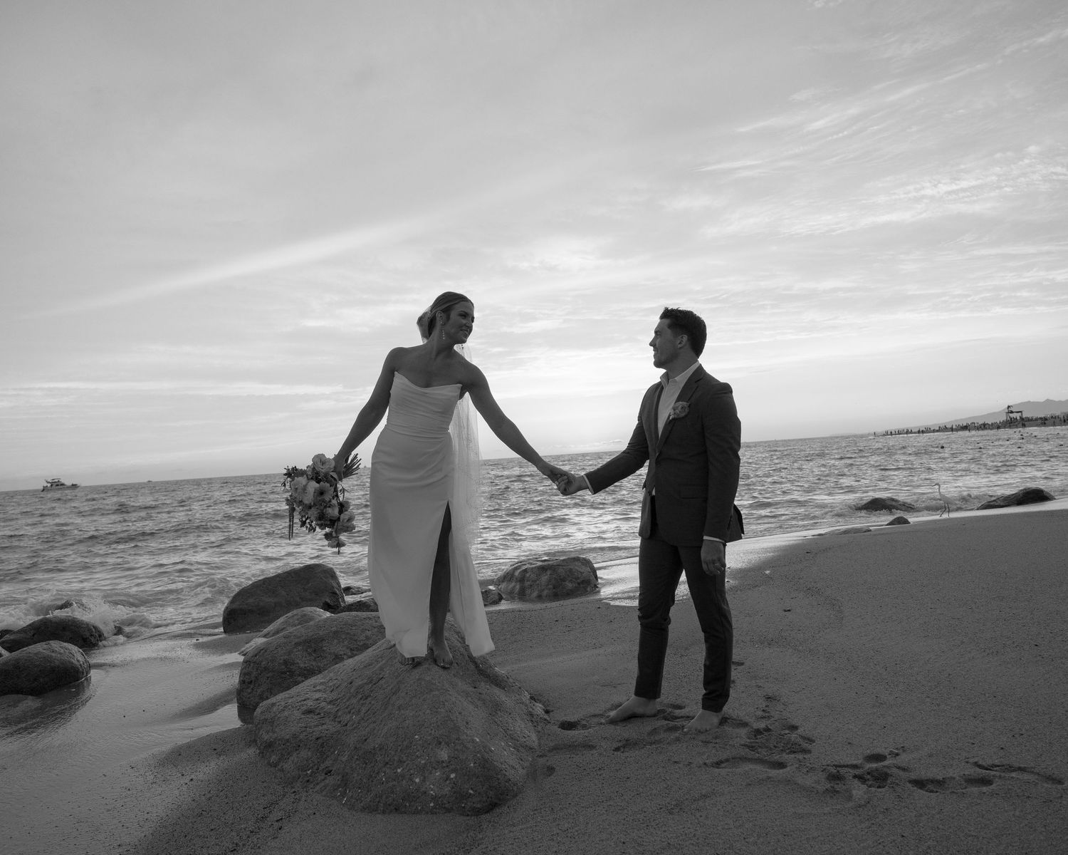 Black and white photo of bride and groom holding hands on a rocky beach at sunset.